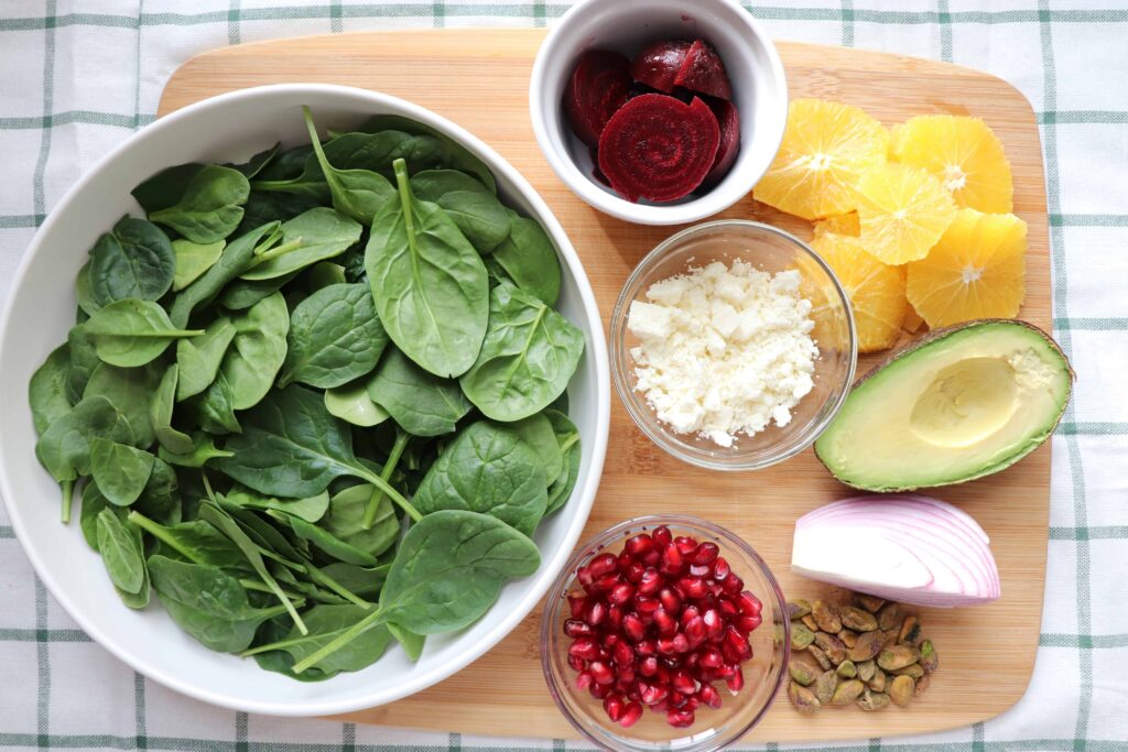 wooden board with fresh spinach, cooked beets, red onions, pomegranate, sliced orange, avocado, feta and pistachios