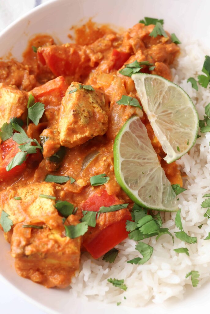 Indian tofu curry with a side of rice garnished with fresh cilantro leaves and lime slices in a white bowl.