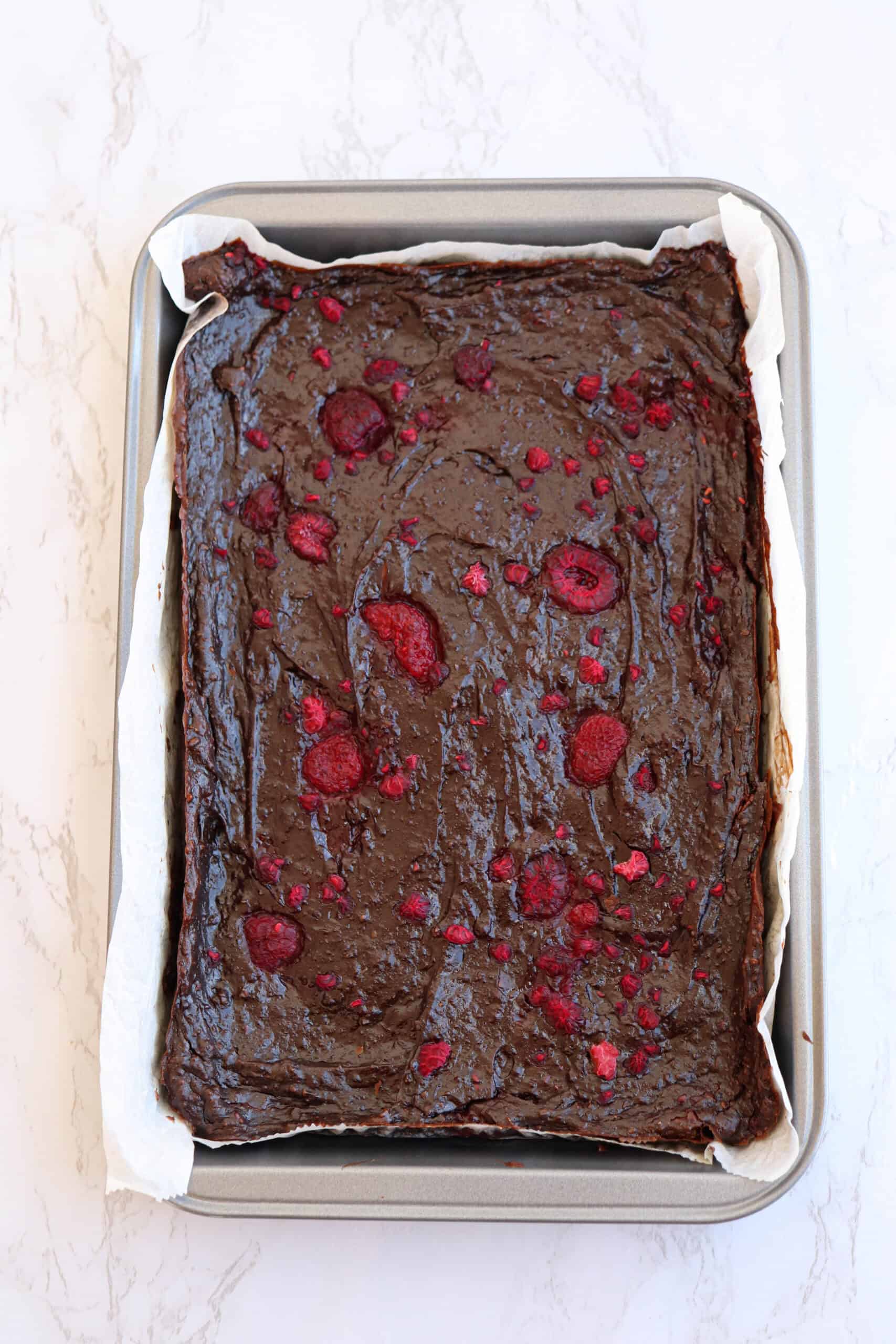 A rectangular baking pan filled with decadent avocado brownies topped with scattered raspberries. The brownies, rich and fudgy, are still in the parchment paper, and the pan rests on a light marble surface.
