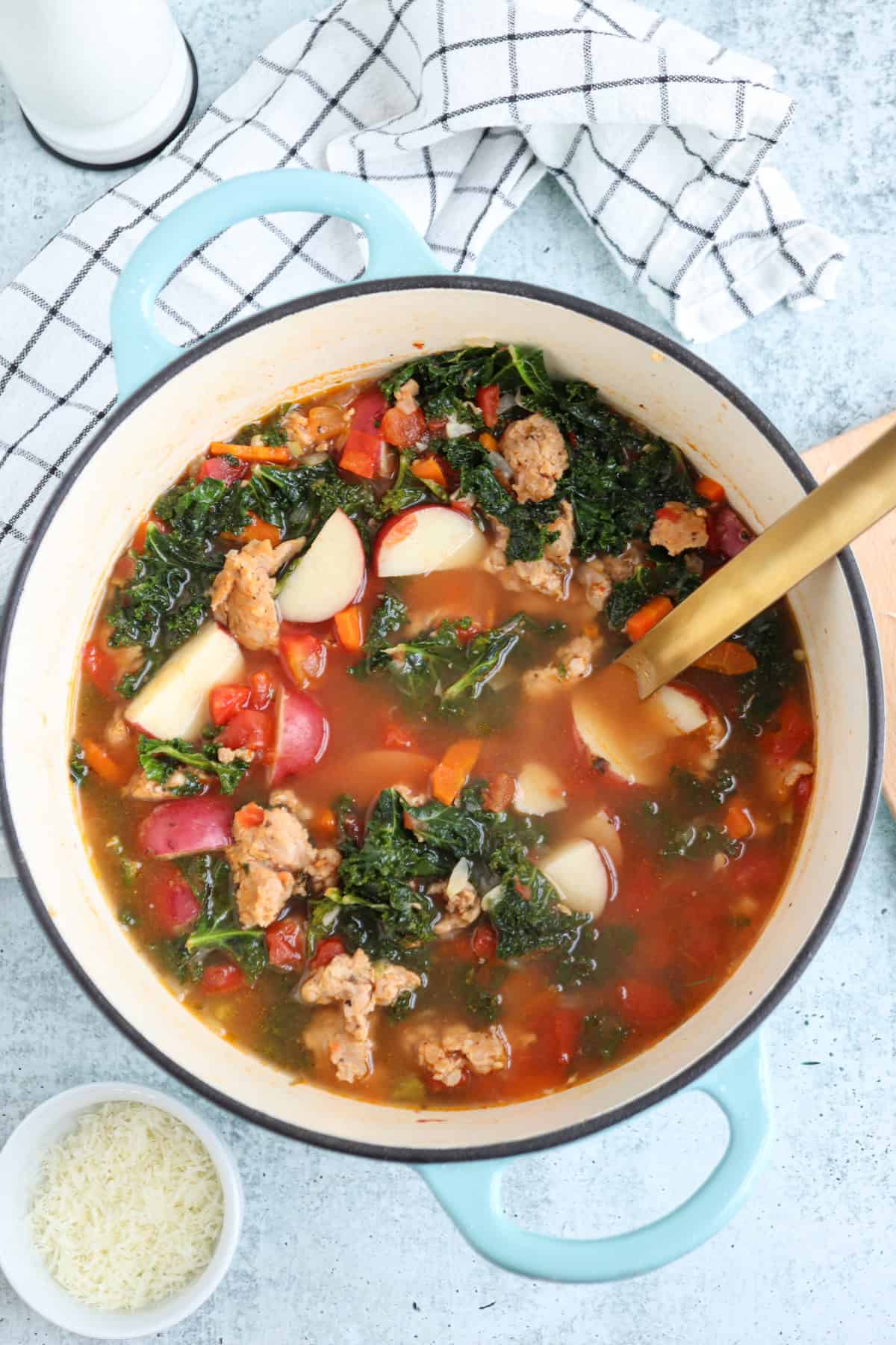 A pot of creamy sausage kale soup brimming with chunks of savory sausage, red potatoes, and diced tomatoes in a rich broth. A wooden spoon rests in the pot. Nearby, a small bowl of grated cheese and a checkered cloth sit on a light blue surface.