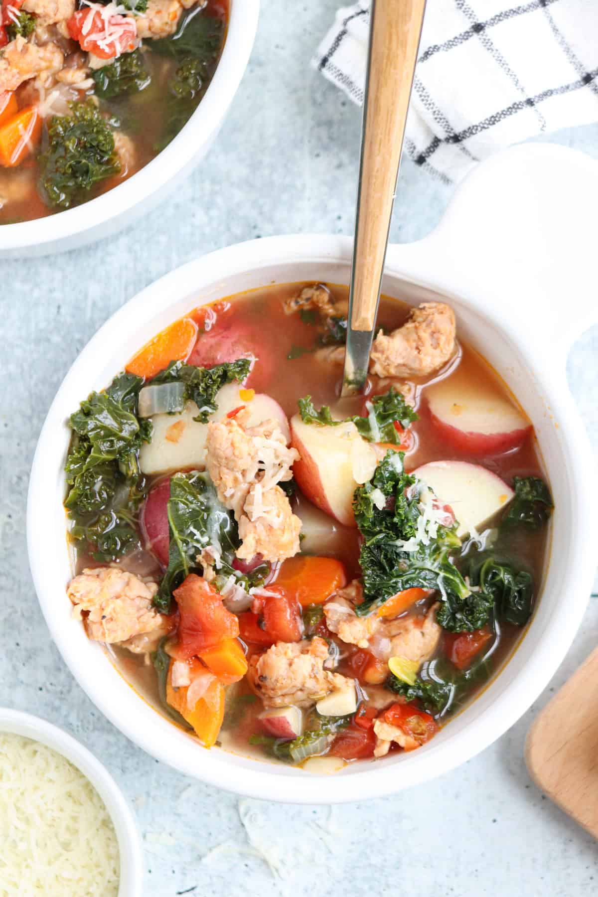 A bowl of creamy sausage kale soup, brimming with chunks of sausage, kale, red potatoes, carrots, and tomatoes in a flavorful broth. A spoon rests in the bowl, with shredded cheese and a napkin nearby.