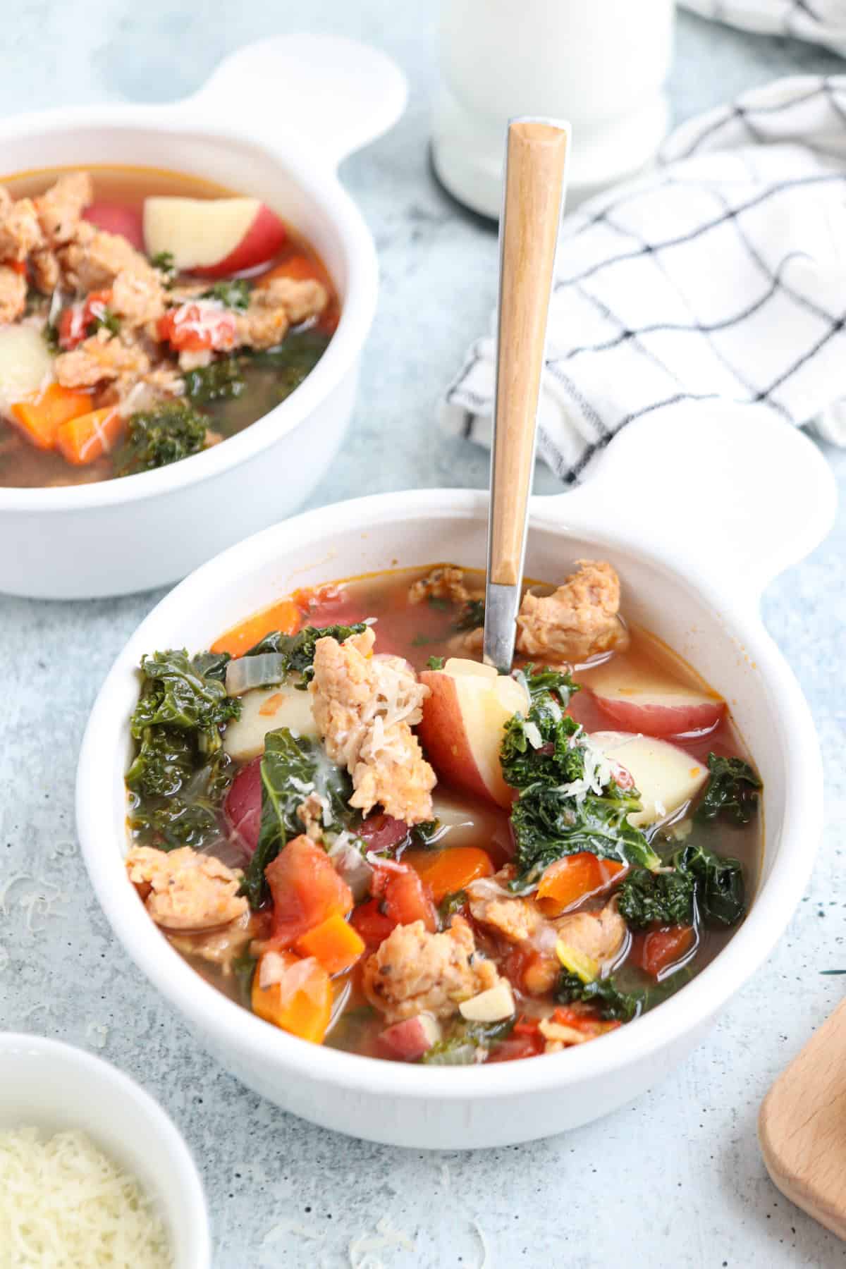 Two bowls of creamy sausage kale soup brimming with chunks of potatoes, carrots, and tomatoes. A spoon rests in one bowl. A checkered napkin and a small dish of grated cheese are nearby on a light gray surface.