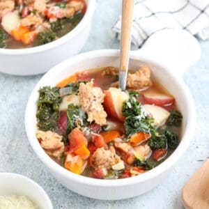 A bowl of creamy sausage kale soup filled with chopped vegetables, sausage pieces, kale, and chunks of potato. The soup is served in a white bowl with a spoon. Another bowl and a checkered cloth are visible in the background.