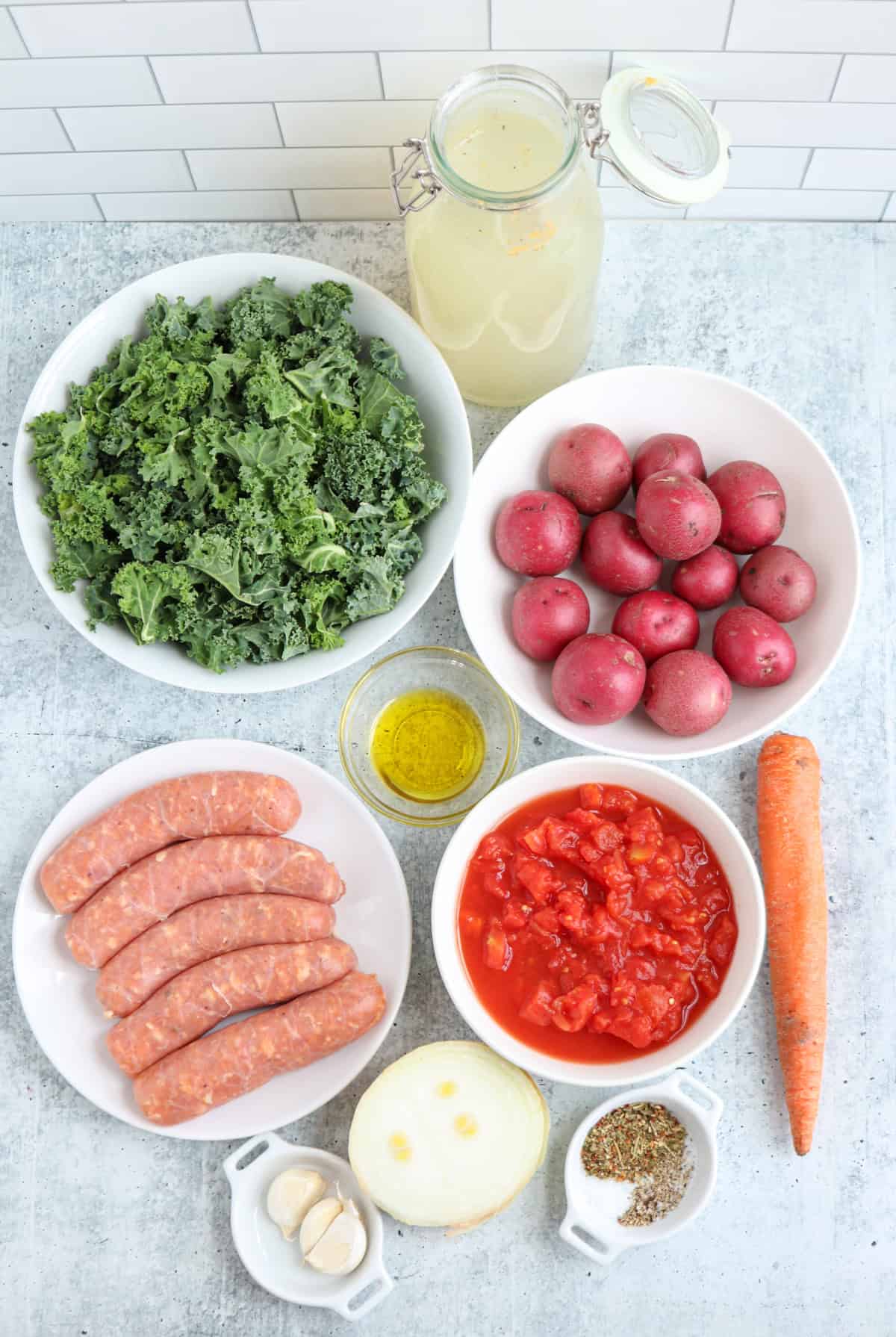Bowls of ingredients for creamy sausage kale soup on a countertop: fresh kale, red potatoes, Italian sausages, diced tomatoes, a carrot, an onion, garlic cloves, a jar of broth, olive oil, and a small dish of herbs.