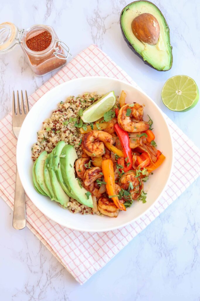 a bowl of shrimp, peppers, onions over quinoa with cilantro and sliced avocado and lime