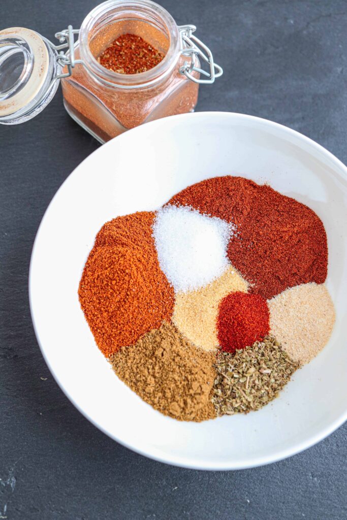 a close up of taco seasoning spices in a white bowl on black background with a small spice jar on top