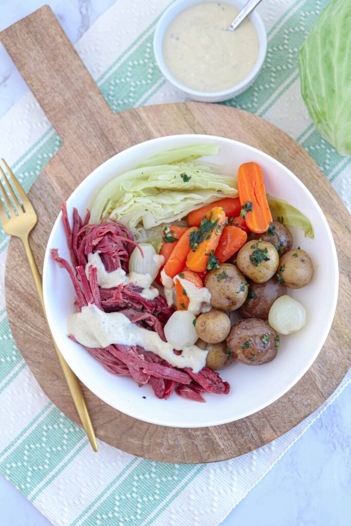 a white plate with corned beef and cabbage, carrots, baby potatoes and horseradish cream on a wooden board