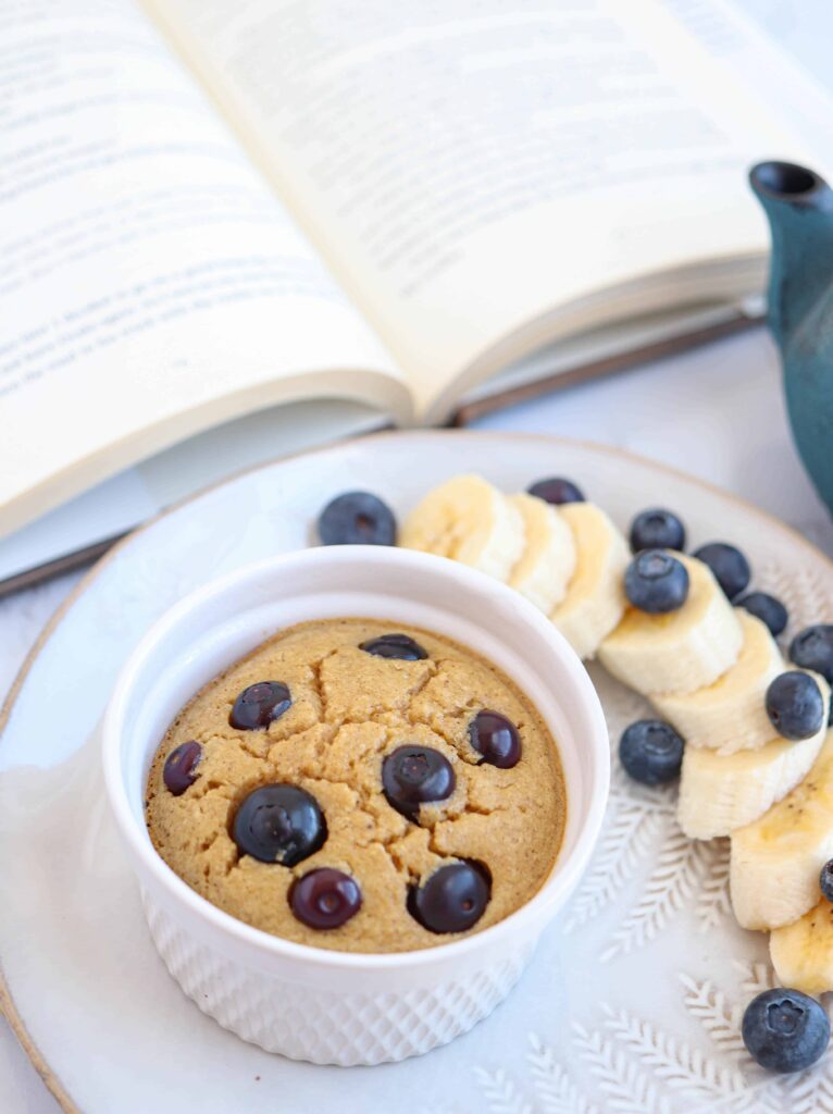a ramekin with banana baked oats with blueberries on top on a plate with a side of fruit and a book in the background