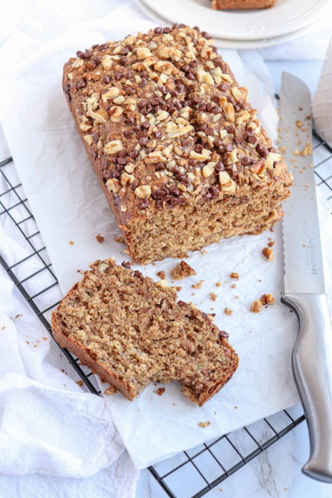 a loaf of banana bread with one slice cut off and a bite taken out ob white paper with a silver bread knife to the side.