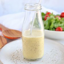A glass bottle filled with Creamy Greek Salad Dressing and a spoon inside sits on a white plate sprinkled with seasoning. In the background, a bowl of fresh greens and cherry tomatoes adds vibrant color to the scene.