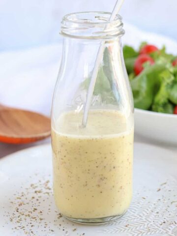 A glass bottle filled with Creamy Greek Salad Dressing and a spoon inside sits on a white plate sprinkled with seasoning. In the background, a bowl of fresh greens and cherry tomatoes adds vibrant color to the scene.