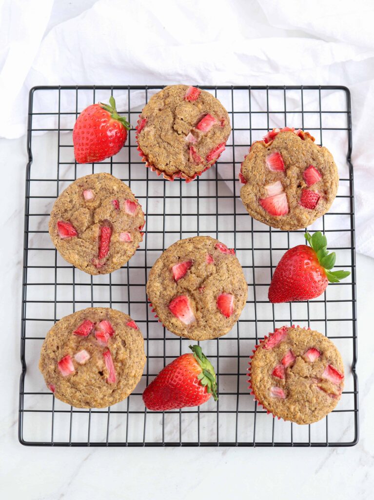 a black cooling rack with strawberry banana muffins in red muffin liners and 3 strawberries around.