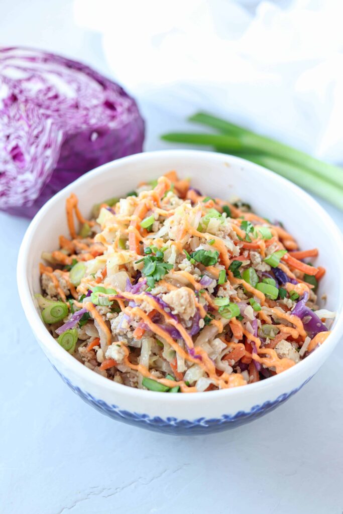 white and clue bowl with meat, veggies, sriracha mayo and cilantro on light gray surface.