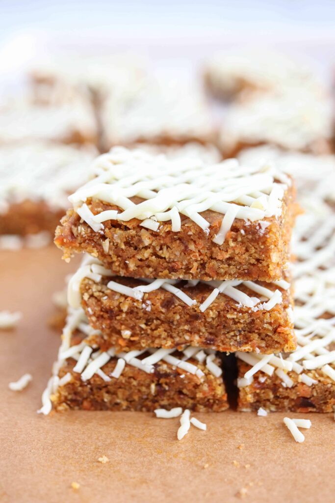 a stack of 3 carrot cake bars wth white chocolate drizzle on brown paper