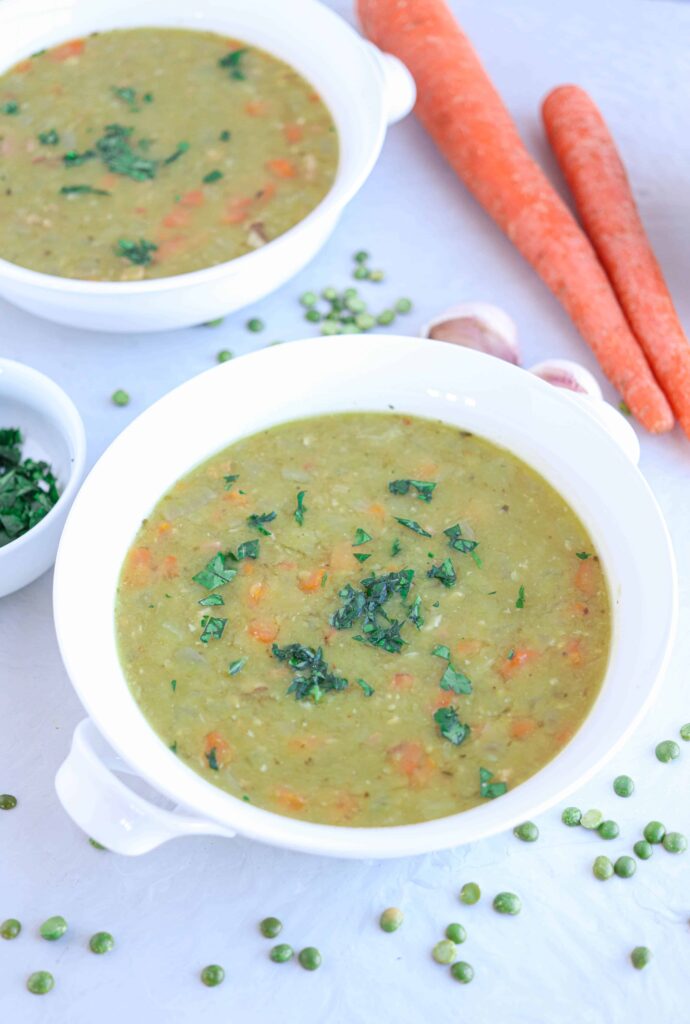 two white bowls with split pea and ham soup on gray countertop with carrots, garlic cloves and dried peas around.