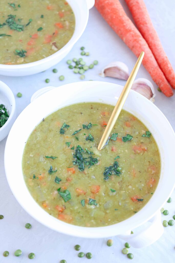 a white bowl with split pea and ham soup on gray background with dried peas, garlic cloves and carrots around 
