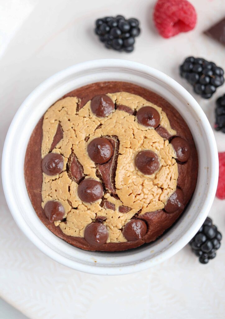 one white ramekin with peanut butter chocolate baked oats on a light colored plate with raspberries and blackberries around.