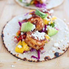 A corn tortilla topped with grilled shrimp, diced avocado, mango salsa, shredded purple cabbage, and crumbled cheese—these Shrimp Tacos with Mango Salsa are served fresh on a wooden surface.
