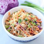 A bowl filled with a colorful, inside out Egg roll-inspired coleslaw featuring shredded red cabbage, carrots, green onions, and chopped herbs. A halved cabbage and green onions are in the background.