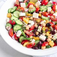 A bowl of Greek Chickpea Salad with cherry tomatoes, cucumbers, yellow bell peppers, red onions, olives, chickpeas, and feta cheese, garnished with chopped herbs.