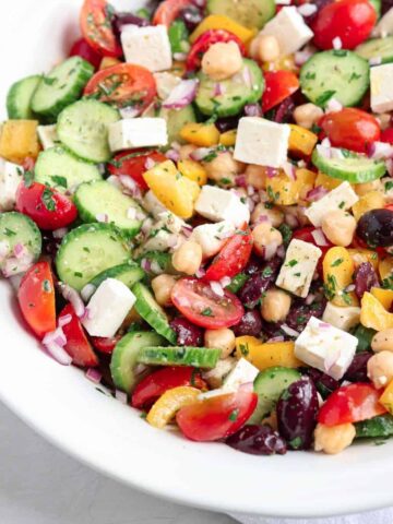 A bowl of Greek Chickpea Salad with cherry tomatoes, cucumbers, yellow bell peppers, red onions, olives, chickpeas, and feta cheese, garnished with chopped herbs.