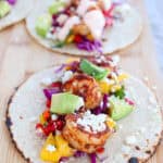 A close-up of two delectable shrimp tacos on a wooden board, bursting with flavor. The front taco is packed with grilled shrimp, diced avocado, mango salsa, red cabbage, chopped tomatoes, and crumbled cheese. The background taco mirrors this vibrant ensemble.
