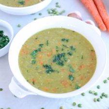 A white bowl filled with Instant Pot Split Pea Soup with ham, garnished with chopped herbs. Carrot pieces are visible in the soup. Carrots, garlic, split peas, and a bowl of parsley are in the background on a light surface.