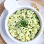 A white bowl filled with creamy Egg Salad with avocado, garnished with chopped fresh dill, sits on a white plate with a fork beside it. The salad features chopped eggs, avocado, and herbs mixed with a creamy dressing.