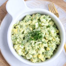 A white bowl filled with creamy Egg Salad with avocado, garnished with chopped fresh dill, sits on a white plate with a fork beside it. The salad features chopped eggs, avocado, and herbs mixed with a creamy dressing.