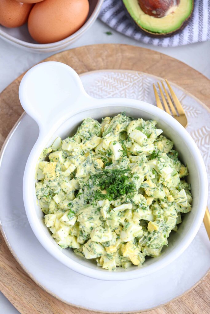 a white bowl with herbed avocado egg salad on a light colored plate on a wooden board with a gold fork next to the bowl