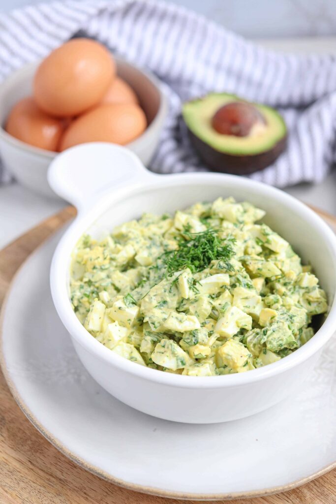 a white bowl with herbed egg avocado salad on a light colored plate on a wooden board. In the background there are eggs in a bowl, half an avocado and a white and gray striped tea towel