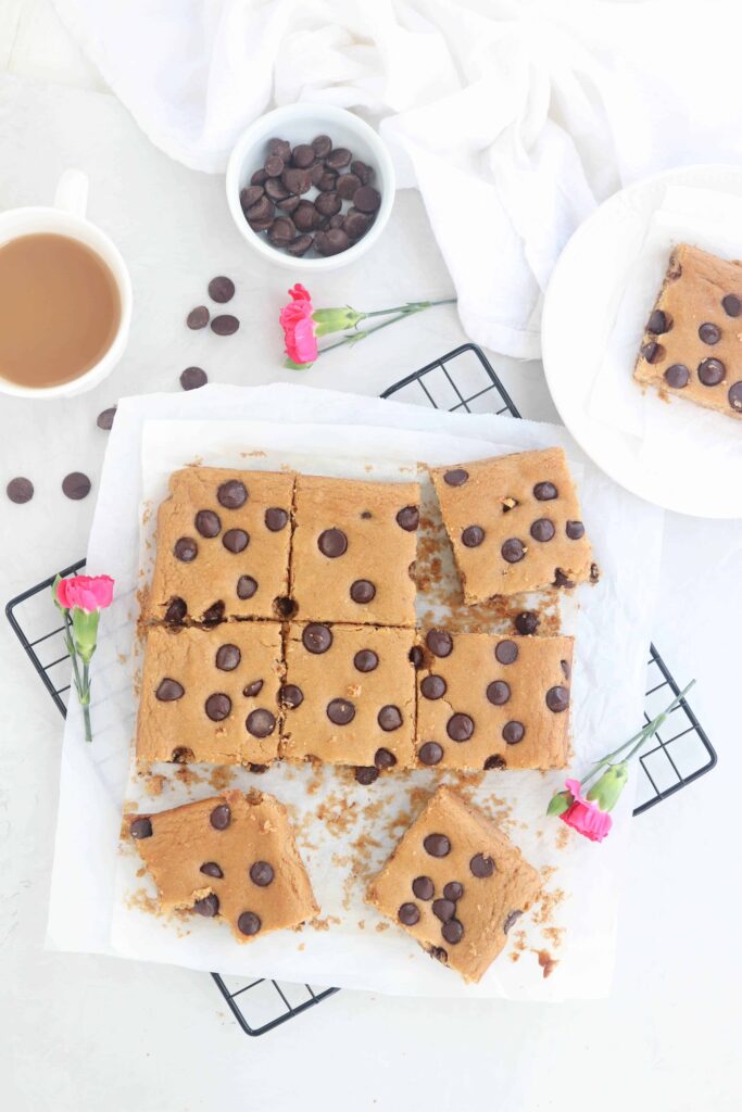 chickpea blondies with chocolate chips cut into squares on white parchment paper on a black cooling rack on light gray surface with 3 pink carnations, chocolate chips and a white cup of tea around 