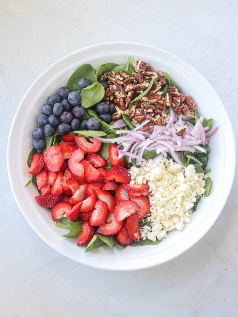 a big white bowl with baby spinach, strawberries, blueberries, red onions, pecans, feta cheese on light gray surface