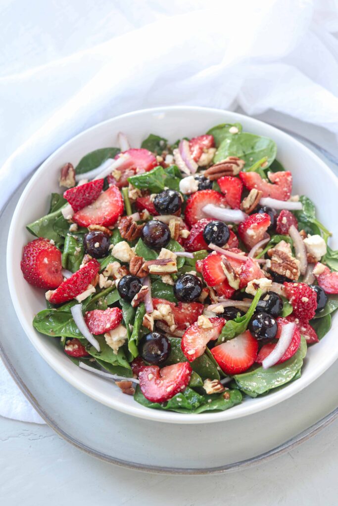 spinach, strawberry salad with blueberries, red onions, feta cheese and pecans on a white bowl on white surface.