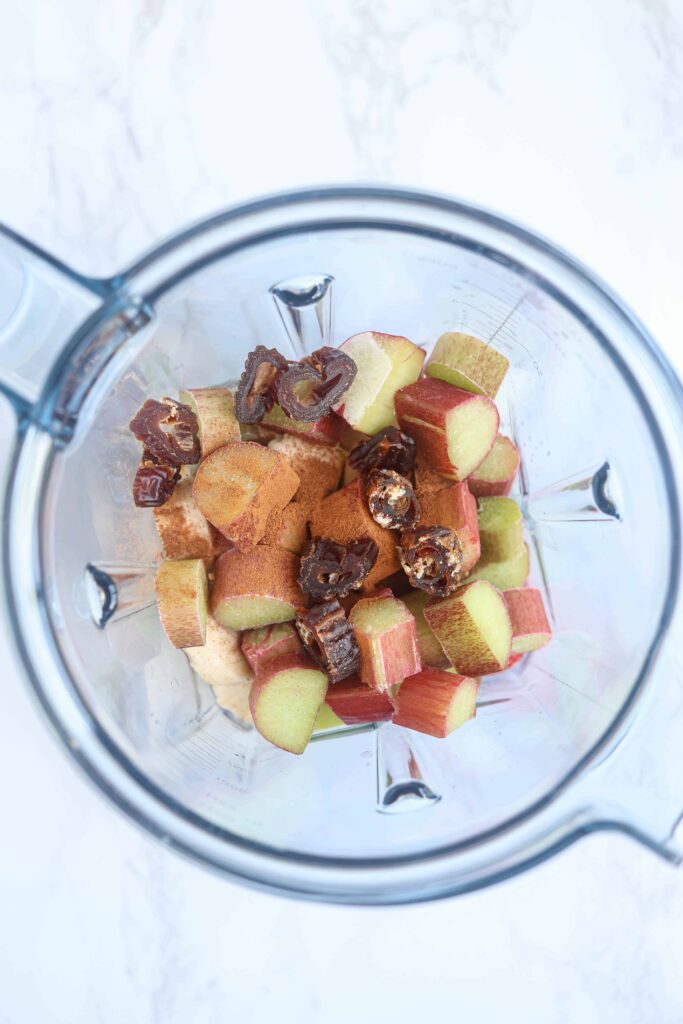 an overhead view of a bowl of a Vitamix blender with rhubarb, dates, cinnamon on white marble surface.