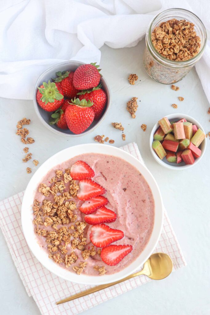 a white bowl with strawberry rhubarb smoothie bowl topped with granola and fresh strawberry slices on a folded white and pink tea towel on light gray surface. Around there are dishes with strawberries, rhubarb and granola, white cloth and a golden spoon