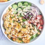 A bowl of salad with Cauliflower Shawarma, sliced cucumbers, cherry tomatoes, red onions, chickpeas, olives, lettuce, and a drizzle of creamy dressing, garnished with black sesame seeds.
