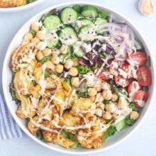 A bowl of salad with Cauliflower Shawarma, sliced cucumbers, cherry tomatoes, red onions, chickpeas, olives, lettuce, and a drizzle of creamy dressing, garnished with black sesame seeds.