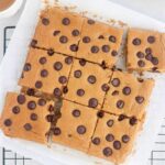 A batch of protein blondies cut into squares on parchment paper, some pieces slightly separated, with visible chocolate chips on top. The bars rest on a cooling rack.