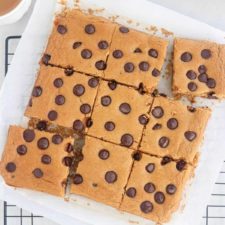 A batch of protein blondies cut into squares on parchment paper, some pieces slightly separated, with visible chocolate chips on top. The bars rest on a cooling rack.