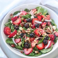 A bowl of strawberry spinach salad with feta, topped with blueberries, pecans, and red onion, is served on a white plate with a white napkin in the background.