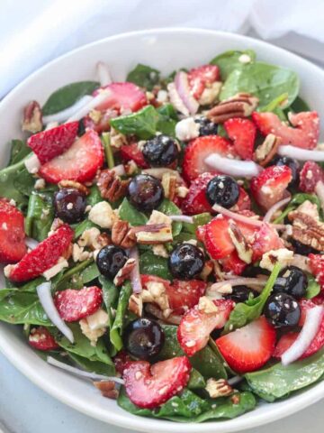 A bowl of strawberry spinach salad with feta, topped with blueberries, pecans, and red onion, is served on a white plate with a white napkin in the background.