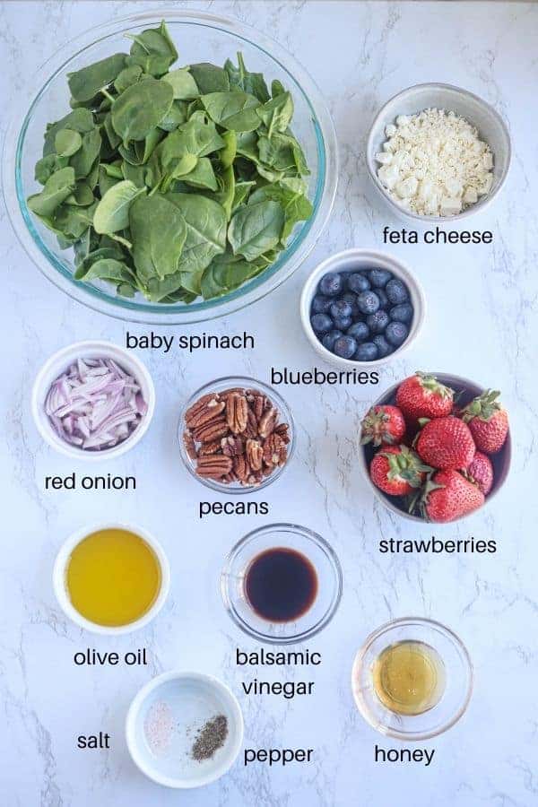 spinach strawberry salad ingredients laid out in bowls on white marble surface with text labels.