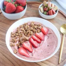 A strawberry rhubarb smoothie bowl topped with granola and sliced strawberries sits on a wooden tray. Nearby are bowls of fresh strawberries and chopped rhubarb, with a gold spoon beside the vibrant smoothie bowl.