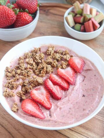 A strawberry rhubarb smoothie bowl topped with granola and sliced strawberries sits on a wooden tray. Nearby are bowls of fresh strawberries and chopped rhubarb, with a gold spoon beside the vibrant smoothie bowl.