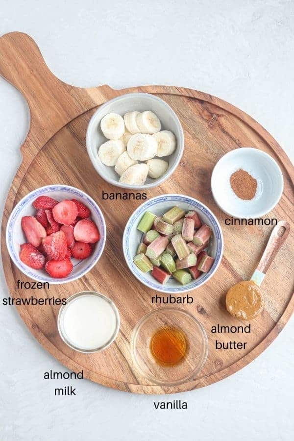 a round wooden board with small round white dishes containing frozen strawberries, rhubarb, bananas, cinnamon, almond milk, vanilla and almond butter on light gray surface.