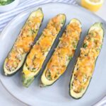 Four zucchini halves on a plate, hollowed out and stuffed with a savory tuna melt mixture of vegetables, then topped with melted cheddar cheese. A lemon half and a striped napkin are in the background.