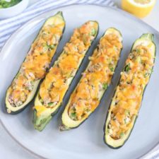 Four zucchini halves on a plate, hollowed out and stuffed with a savory tuna melt mixture of vegetables, then topped with melted cheddar cheese. A lemon half and a striped napkin are in the background.