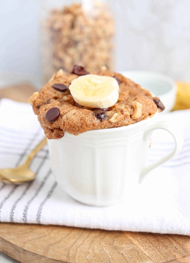 a white mug with banana cake with chocolate chips and a slice of banana on a wooden board on a white napkin with a golden spoon on the left.