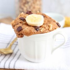 A white mug filled with Banana Cake in a mug, topped with chocolate chips, walnuts, and a banana slice, sits on a striped cloth with a gold spoon nearby.